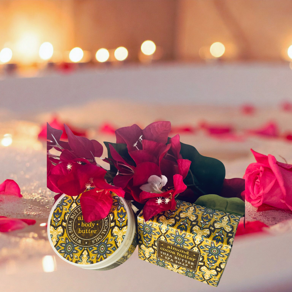 A round tin and matching rectangular box of body butter with ornate gold and teal floral patterns, styled with red flowers and rose petals on a soft, warm, blurred background with bokeh lights.