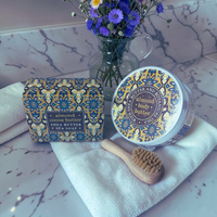 Thumbnail for A jar of almond body butter and its matching box are displayed on a white towel with a wooden brush, set against a marble-patterned background with a small vase of purple flowers.