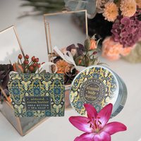 Thumbnail for A styled product shot of almond body butter and shea soap in decorative packaging, arranged with dried flowers, a mirror, and a pink lily on a light surface. The round tin and rectangular box both feature a blue and gold damask pattern with black text reading 'almond body butter' and 'almond cocoa butter shea soap'.