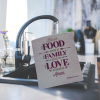 Thumbnail for A rectangular kitchen towel with a printed religious quote hangs from a black faucet in a kitchen sink. The towel reads, 'Bless the FOOD before us the FAMILY beside us the LOVE between us Amen.