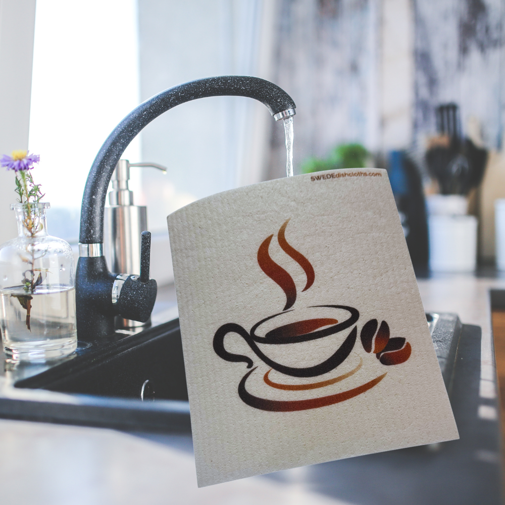 A square kitchen towel with a coffee cup graphic is draped over a modern kitchen sink with water running from a black faucet.