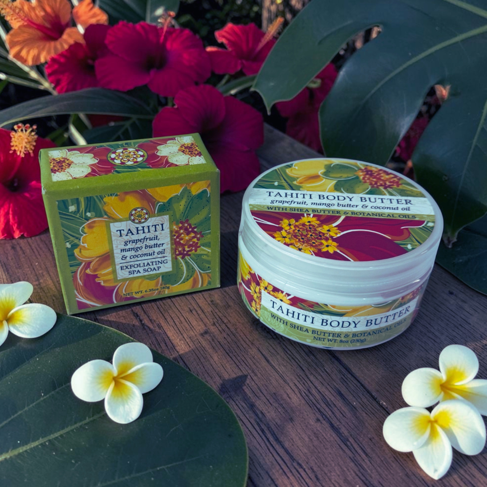 Tahiti Body Butter exfoliating body butter jar and matching box displayed on a wooden surface with tropical flowers and leaves in the background.