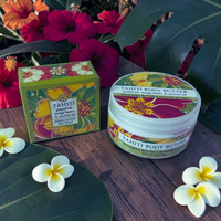 Thumbnail for Tahiti Body Butter exfoliating body butter jar and matching box displayed on a wooden surface with tropical flowers and leaves in the background.