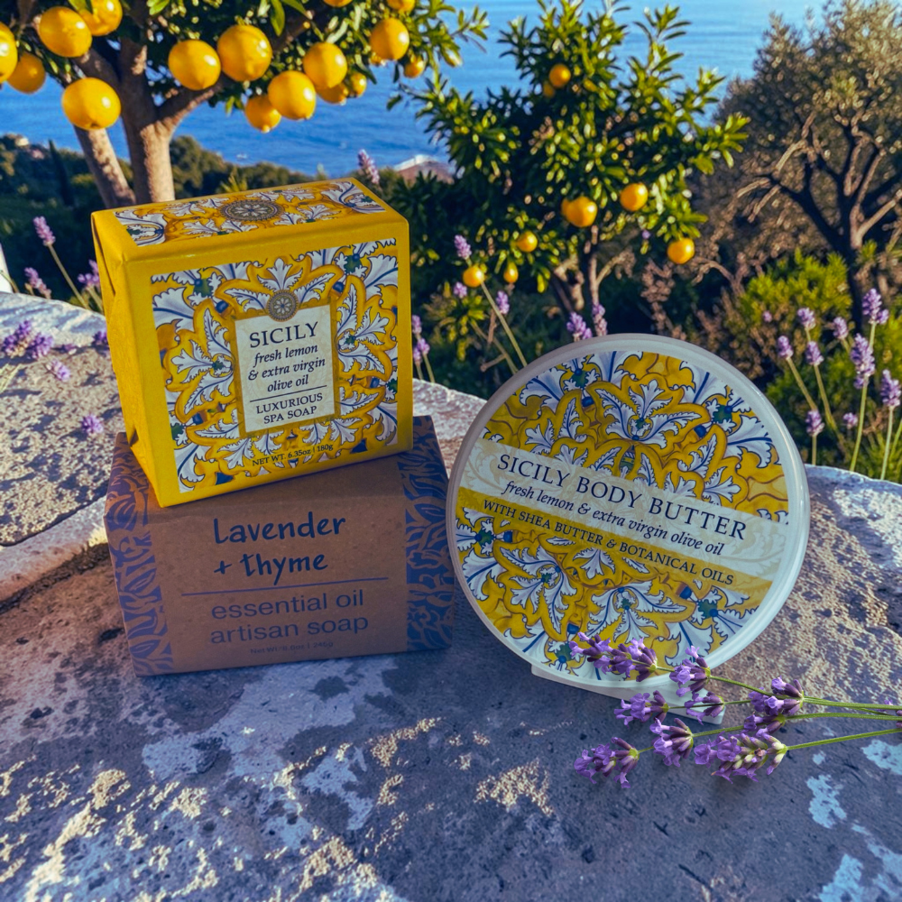 Sicily Body Butter and Sicily Lavender + Thyme artisan soap products displayed outdoors on a stone surface with lemons, lavender, and a sea background. The round body butter tub and the branded yellow-and-white box are shown, along with a smaller brown box for the lavender and thyme soap.