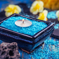 Thumbnail for A square dark glazed ceramic candle holder filled with bright blue salt crystals, with a small lit tealight candle floating in the center. The scene is styled with blue salt crystals, a porous stone piece, and soft yellow flowers in the blurred background.
