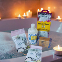 Thumbnail for A bath scene with a bathtub, lit candles, and several self-care items arranged on the tub edge: a bottle labeled Lavender Cleansing Oil, a pink stand-up pouch, a yellow and purple 'Morning Glamour' satin sleep set package, a lavender + rhinestone box, and two white bath salts pouches. No person or animal is present, but this is not a clean isolated studio shot; it is a styled real environment, so subject_type is Lifestyle.