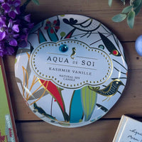 Thumbnail for Decorative candle tin with 'Aqua de Soi' branding on a wooden surface with flowers and books.