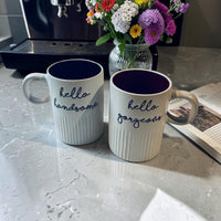 Thumbnail for Two white ribbed ceramic mugs with dark purple interiors and navy script text reading 'hello handsome' and 'hello gorgeous' on a gray marble countertop, with a vase of flowers and an open magazine in the background.