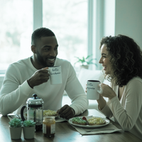 Thumbnail for A man and a woman sitting at a table having breakfast, each holding a white ceramic mug with the text 'hello breakfast'.