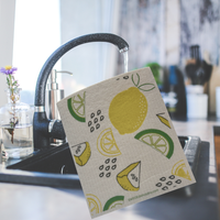 Thumbnail for A square kitchen towel with a lemon and lime slice pattern hangs from a black faucet in a kitchen sink. The towel has a light beige background with yellow lemons, green lime wedges, and black dotted details.