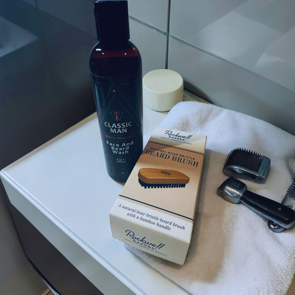 Men's grooming products including a face and body wash bottle, beard brush, and packaging on a towel.