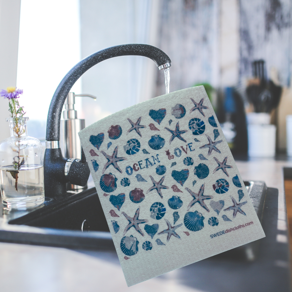 A square kitchen towel with a blue and purple seashell and starfish pattern hanging from a running faucet in a kitchen sink. The towel features the words 'OCEAN! LOVE' and the brand name 'sweeDESweetsCo.