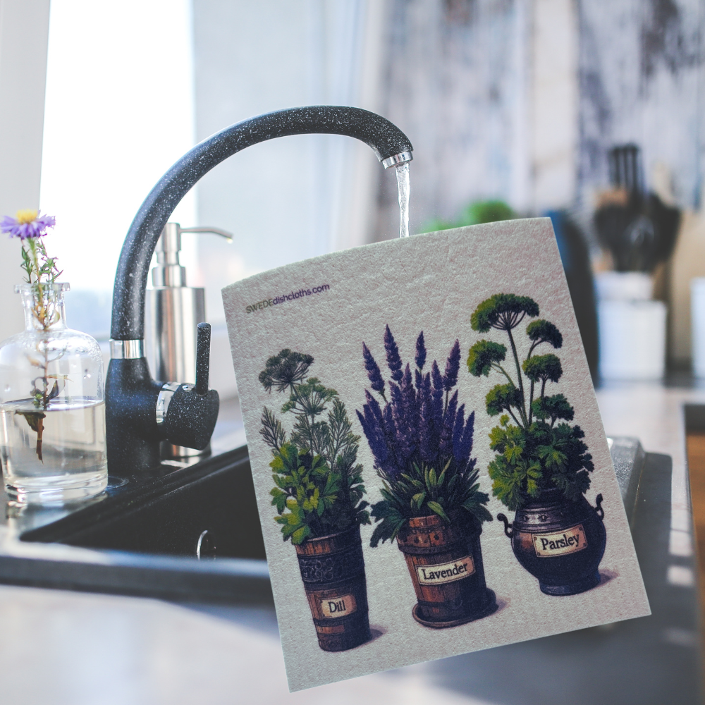 A square kitchen towel or coaster featuring an illustration of three potted herbs (dill, lavender, parsley) is held in front of a running faucet in a kitchen sink. The towel has a textured off-white background and is shown in a real-world kitchen environment.