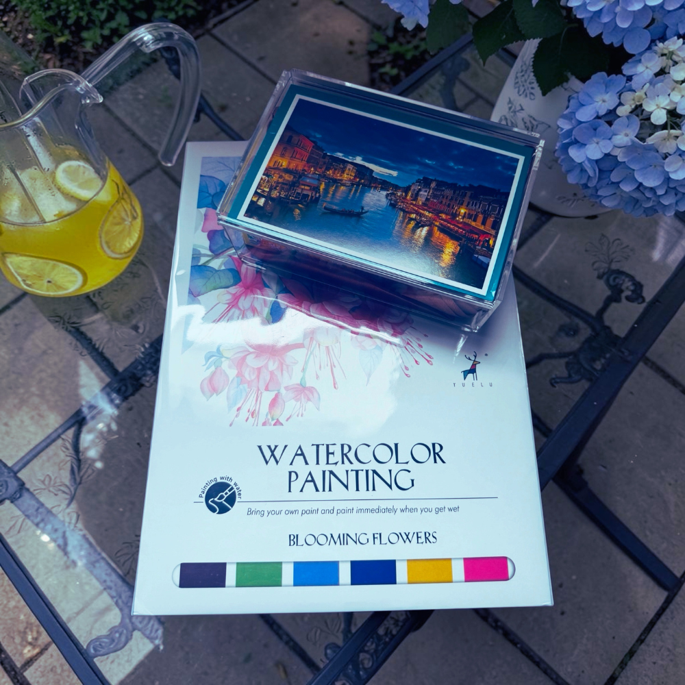 Artistic set with watercolor painting book and cityscape card on a table with a drink and flowers.