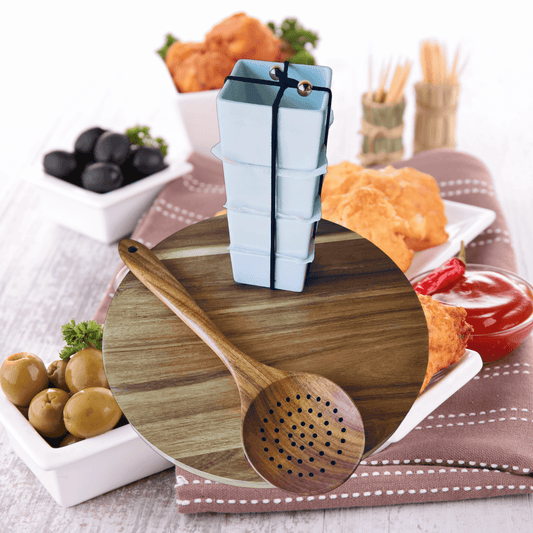 Acacia wood charcuterie board with assorted snacks, including olives, crackers, and condiments, along with a cheese slicer on a rustic table setting.