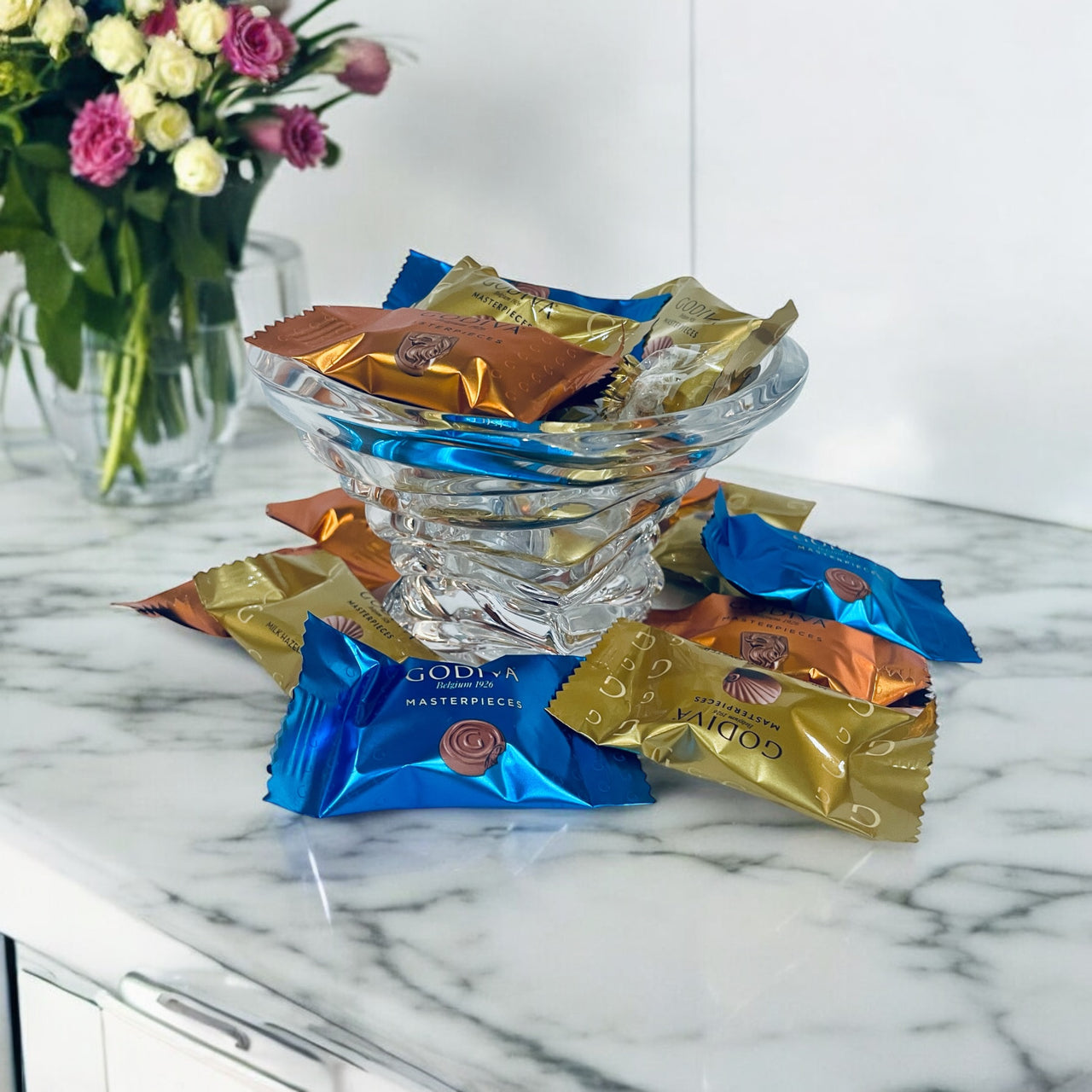 Godiva chocolate bars in colorful wrappers on a marble surface with a vase of flowers in the background.