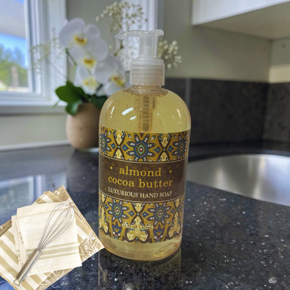 A pump bottle of luxurious hand soap labeled 'almond cocoa butter' sits on a dark kitchen countertop, with a matching soap dish and whisk nearby and white flowers in the background.