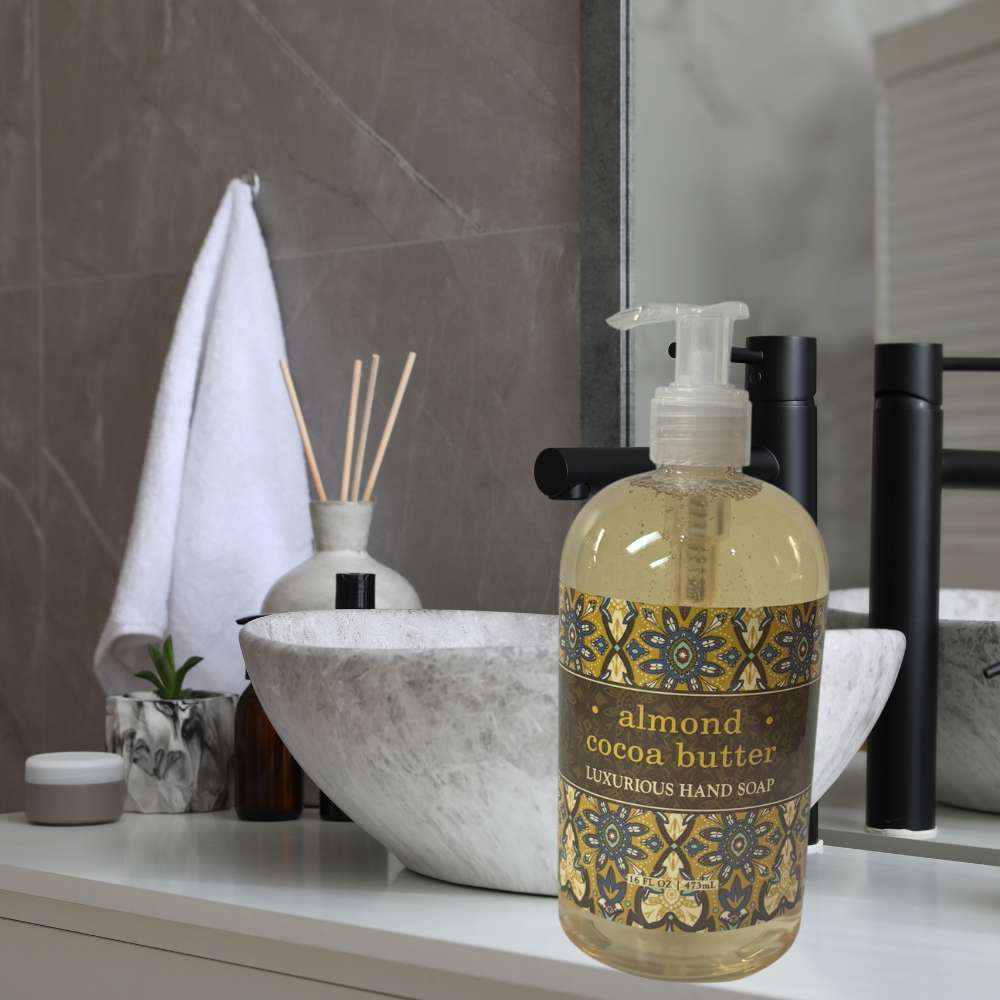 A bottle of almond cocoa butter luxurious hand soap with a gold and blue patterned label, placed on a bathroom counter next to a marble bowl, black faucet, and a white towel.