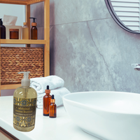 Thumbnail for A bottle of almond cocoa butter hand soap with a white pump and ornate gold label sits on a white bathroom counter next to folded white towels and amber dropper bottles, with a wooden shelf and gray tiled wall in the background.
