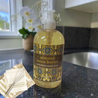 Thumbnail for A pump bottle of luxurious hand soap labeled 'almond cocoa butter' sits on a dark kitchen countertop, with a matching soap dish and whisk nearby and white flowers in the background.