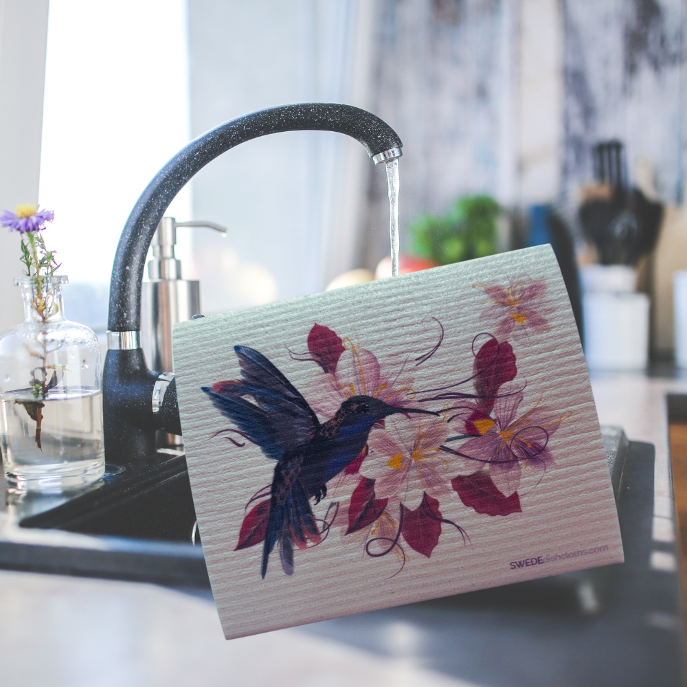 A textured dish towel with a watercolor design of a hummingbird and flowers, held in front of a running faucet in a kitchen setting.