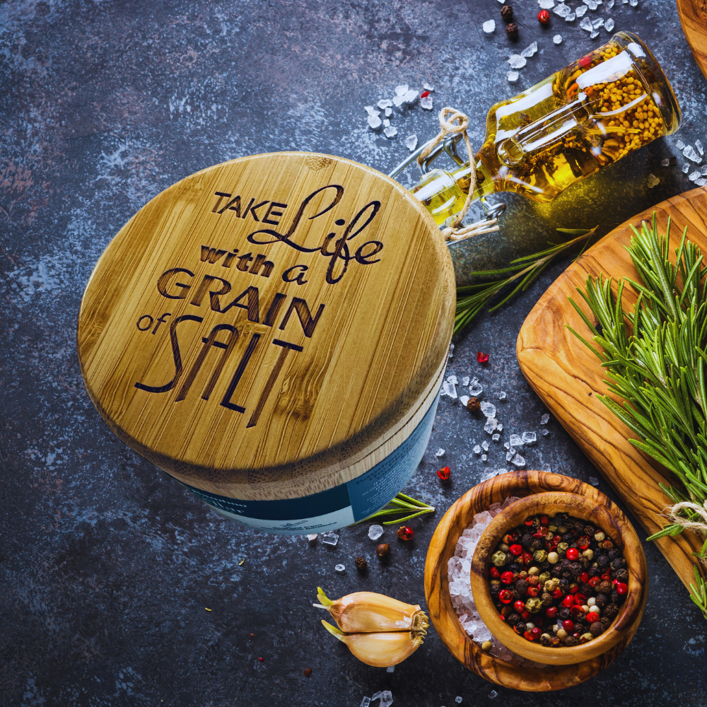 Wooden salt container with 'Take Life with a Grain of Salt' text, surrounded by spices and herbs on a dark surface.