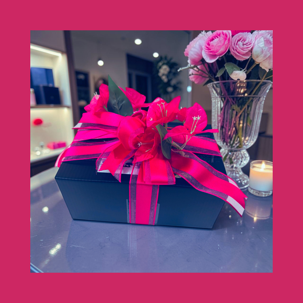 A black rectangular gift box tied with a bright pink ribbon and decorated with artificial pink flowers, placed on a reflective tabletop in an indoor setting with a vase of pink roses and a candle in the background.