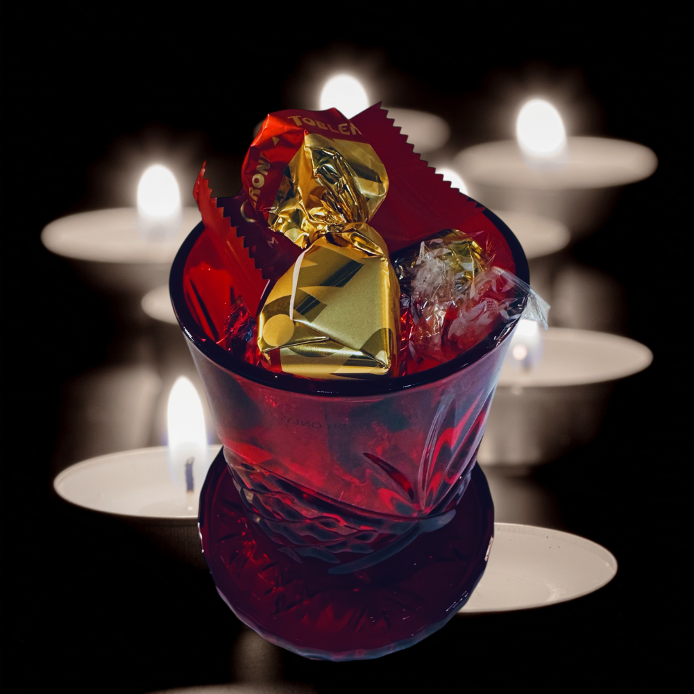 Red glass bowl with gold and red candies against a dark background with blurred candles