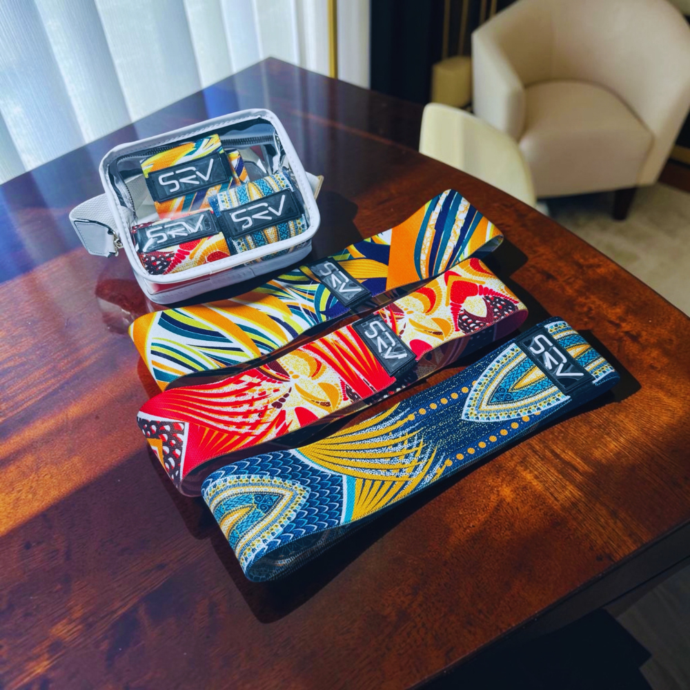 Three colorful patterned belt-like accessories with visible SRY branding are arranged on a wooden table, with an open matching zip case above them in a real indoor room setting.