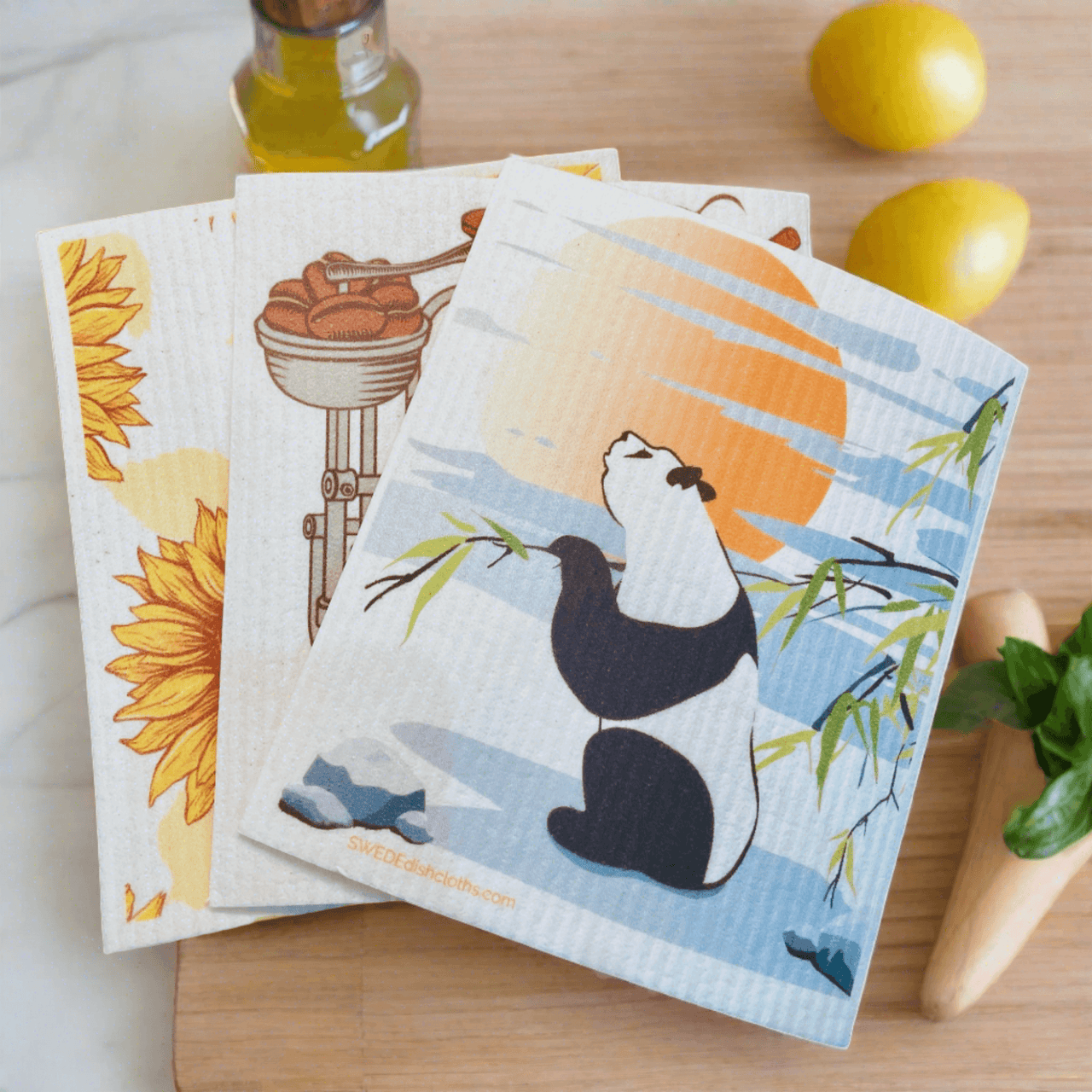 Illustrated dishcloths with panda, sunflower, and kitchen motifs on wooden surface with lemons and bottle of oil.