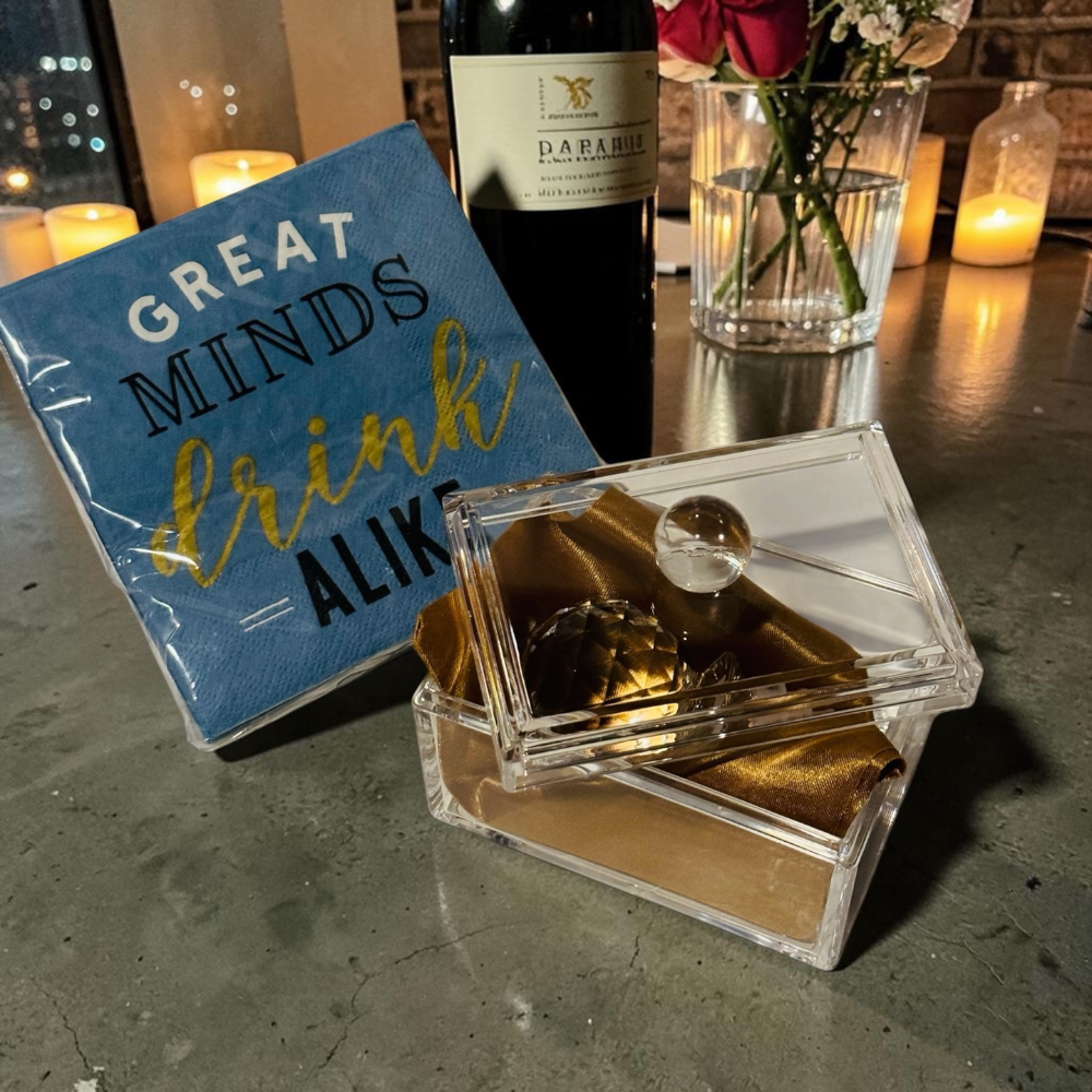 Decorative box with gold interior, blue card with 'Great Minds Drink Aliki' text, and wine bottle on a table.